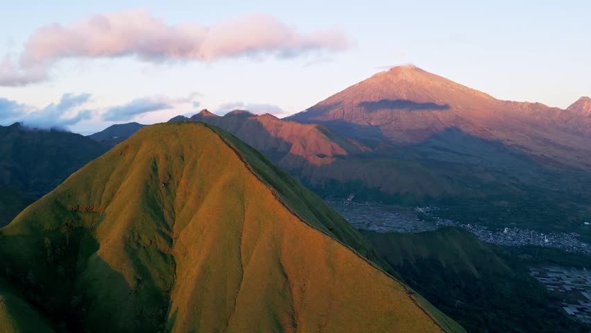 Golden sunrise light illuminating grassy mountain ridge leading toward summit above Sembalun village with Mount Rinjani dominating volcanic landscape in Lombok Indonesia, rising drone footage