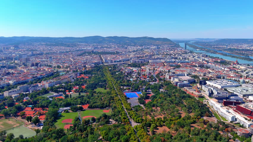 4K aerial drone footage over Vienna, Austria. Prater park with tree-lined Hauptallee, Riesenrad, Donaukanal, WU Wien campus, and Danube River stretching to the Wienerwald.