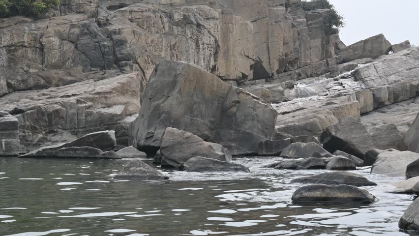 A rocky waterfall edge. Water from a small stream cascades down, creating turbulence in the pool below. Large, layered rock formations and boulders meet the calm, rippling water. The rocks are grey and beige, featuring distinct angular fissures.
