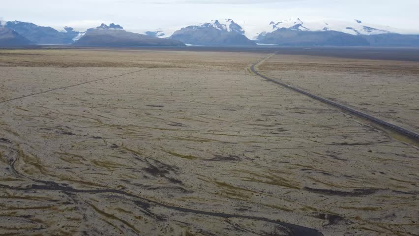 Aerial View of Road Through Icelandic Moss Fields and Snowy Mountains - High-angle drone shot of a lonely road traversing vast mossy lava plains toward distant glaciers and snow-capped peaks under an overcast sky.