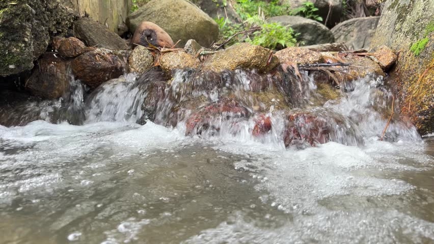 Slow-motion close-up of a small, bubbling waterfall cascading over smooth river rocks in a lush forest stream.