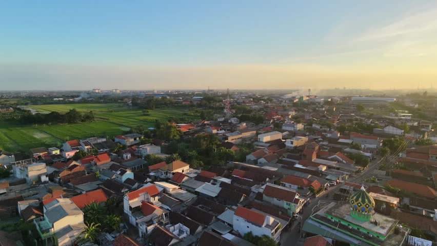 Slow aerial drone tracking shot of a densely populated suburban residential area and green rice fields during golden hour sunset in Sidoarjo, East Java, Indonesia.