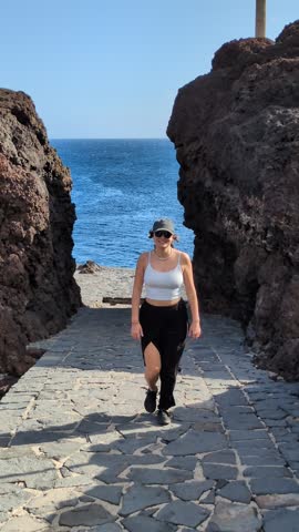 Attractive young woman in casual clothes walking up stone stairs from the blue ocean framed by majestic volcanic rocks in Punta de Teno, Tenerife, Spain, enjoying active summer travel vacation