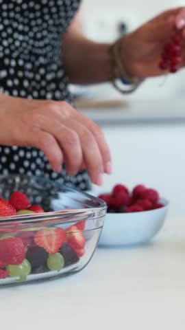 Girl adds raspberries to a bowl of mixed fruits in a bright kitchen. Fresh fruits are ready for a healthy snack.