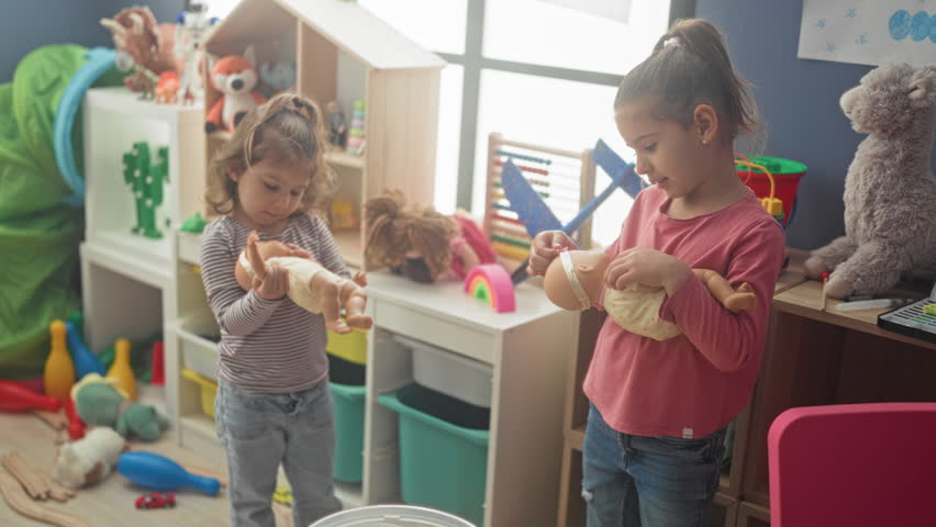 Two girls cradling baby dolls and holding them with both hands in an indoor playroom storage area inside a building surrounded by shelves, toys and bins; tenderness friendship play.