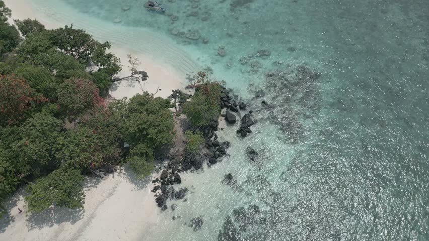 Slow top down aerial hovering over a rocky mangrove islet edge with white sand and crystal clear turquoise shallows near Sebesi Island, Lampung.