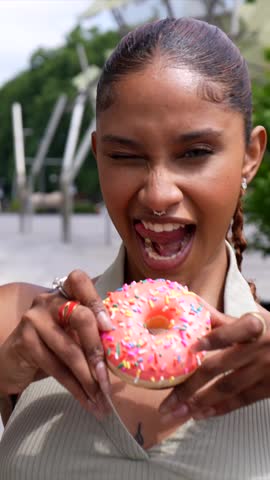 Close up of a happy young woman with a nose ring playfully eating a delicious pink glazed donut with colorful sprinkles, enjoying a sweet treat outdoors on a sunny summer day in the city