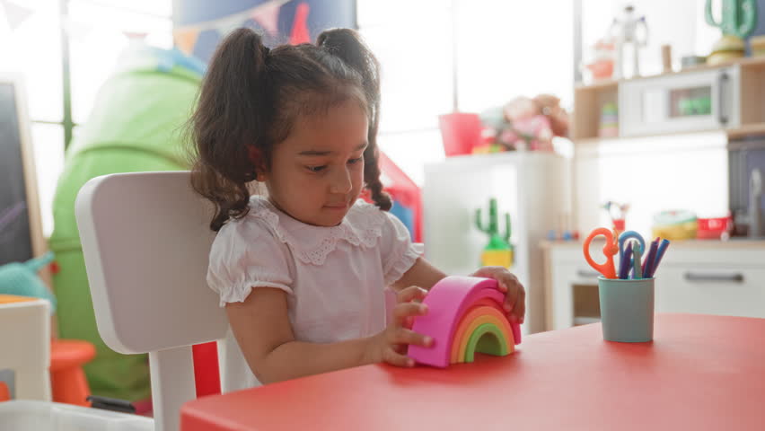 Girl sits at red table playing with rainbow stacking toy, reaches with hand toward colorful cup and scissors in an indoor building; playful curiosity.