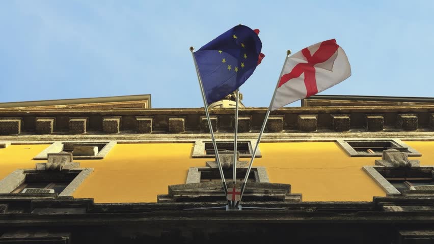 Three prominent flags, the European Union, Italian, and Milanese civic banners, wave gracefully against a clear blue sky from an elegant municipal building. Represents local and national governance, patriotism, and European unity.