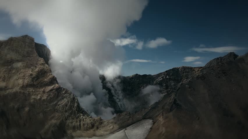 Dramatic drone shot flying over a massive mountain volcano crater emitting thick hot steam and ash into a dark blue sky.