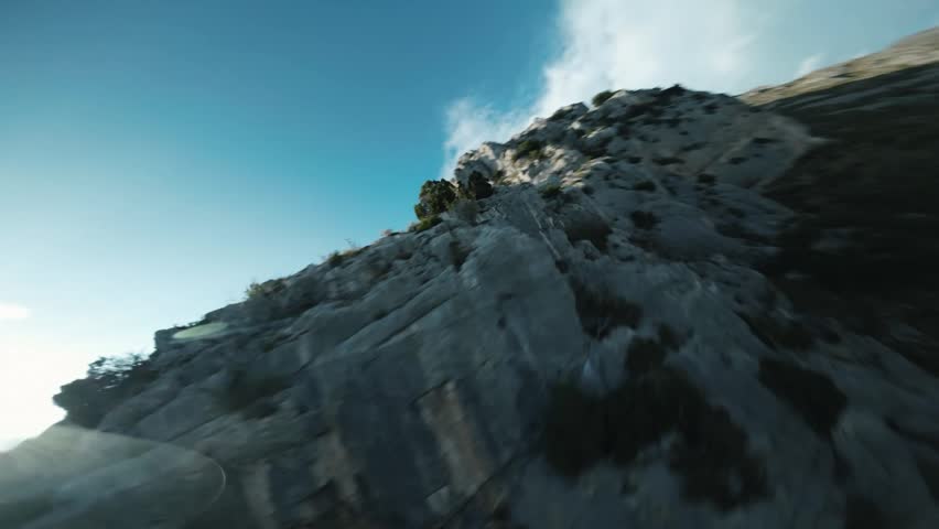 Dynamic first person view drone flying fast over a sharp rocky ridge looking down at a vast valley village landscape.