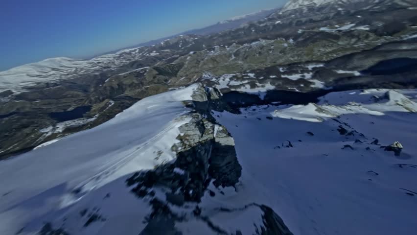 Dynamic first person view racing drone flying low and fast over rugged rocky mountain terrain covered in winter snow.
