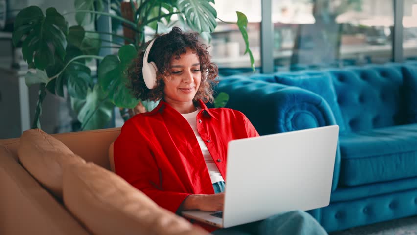Young indian woman sitting relaxed on sofa in homeoffice using laptop computer with headphones for remote freelance work. Female curly hair teenage student girl learning studying online indoor at home