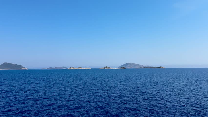 A coastal view of a town nestled between mountains and the sea. The scene features clear blue water and a backdrop of rocky hills