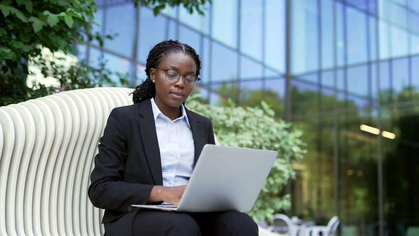 A close-up shot of a professional african american businesswoman in a suit and glasses using her laptop while sitting on a bench. This female employee looks focused as she works on her computer