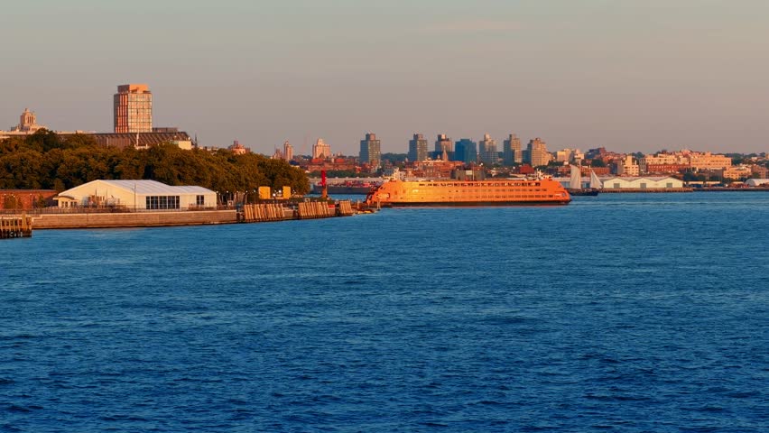 Staten Island Ferry docked at Governors Island terminal. Orange vessel at pier with deep blue harbor water and Brooklyn skyline on the horizon.