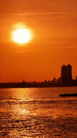 Sunset over New York harbor with city silhouette. Golden sun reflecting on rippled water with buildings in warm orange tones.