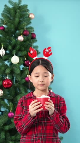 Little asian girl sipping hot cocoa next to a christmas tree