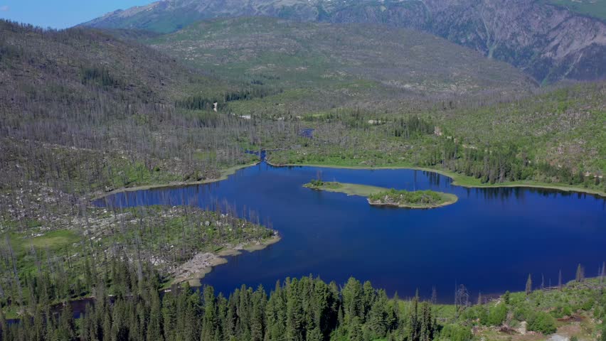 Drone flies sideways, mountain lake with small island surrounded by forest, dead trees in Altai Kazakhstan revealing dramatic valley landscape with strong natural contrast, scenic aerial composition