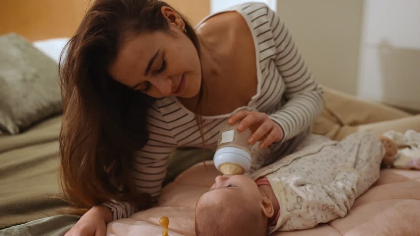 A White mother smiles while holding a bottle to feed her baby as they lie on a bed in a bedroom