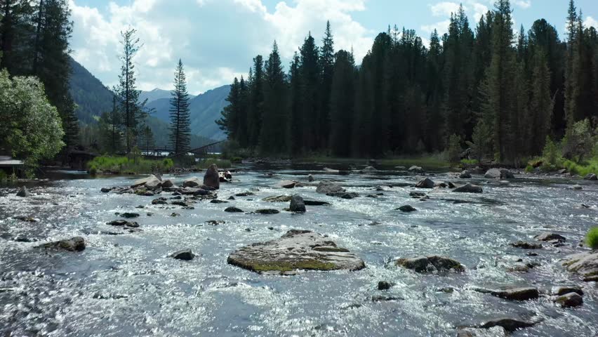 Drone flying upstream along rocky mountain river toward Rakhmanovskoye Lake in Altai, East Kazakhstan. Pine forest, flowing water and distant lake create scenic alpine wilderness landscape.