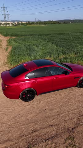 An aerial view of a red sports car driving along a country dirt road through green fields. Vertical Aerial 4K drone footage.
