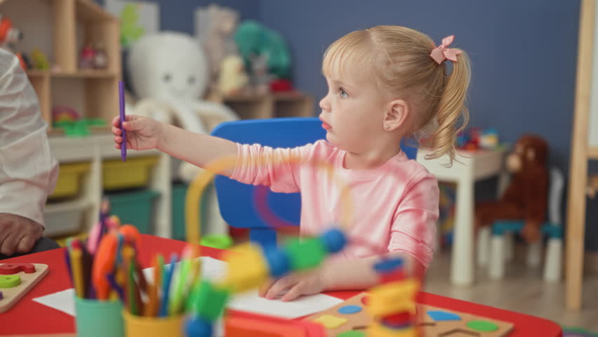Girl reaches for marker handed by a teacher while seated at a craft table with pencils, puzzles and toys in a preschool building; curiosity.