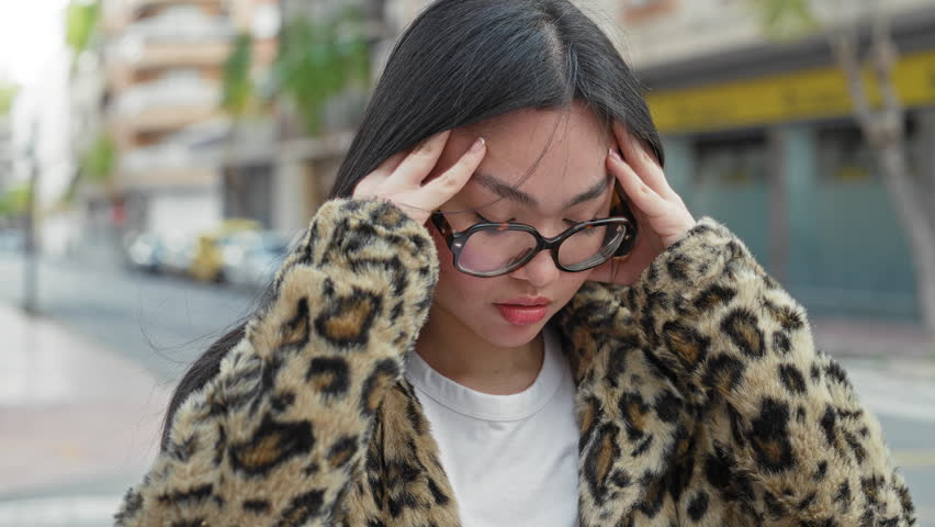 Woman with glasses holding her temples for headache, hands to head on a city street wearing leopard coat; stress, focus.