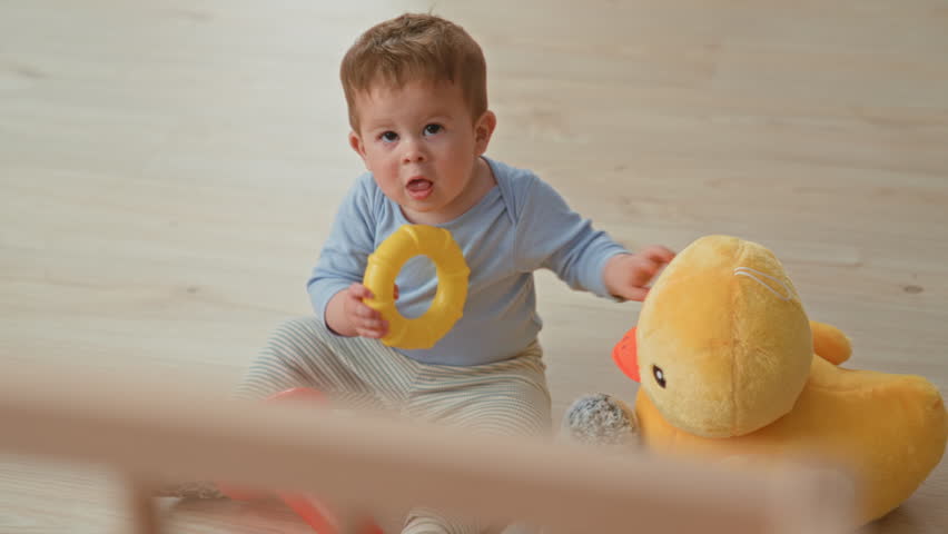 Boy holding a yellow ring toy while sitting on light wood floor, turning his head and gripping the toy beside a large plush duck in building; curiosity.