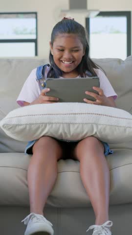 Vertical video: Reacting to tablet child in overalls laughing adjusting grip on home sofa cushion. Device, preteen, denim, scrunchie, couch, daylight, vertical video