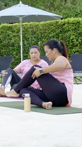 Vertical video: Reacting to partner cue woman in pink doing seated twist on yoga mats peer guiding. Outdoor, poolside, patio, duo, fitness, stretch, vertical video