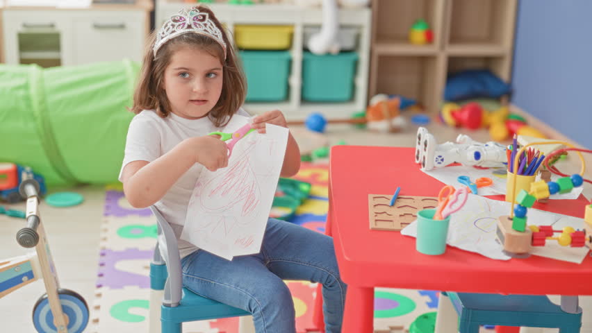 Little girl cutting paper with orange scissors at a red table in building surrounded by toys and art supplies; creative concentration play.