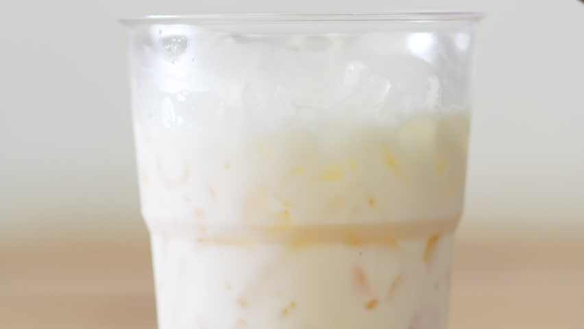 4K Close up of iced coffee latte with swirling dark espresso layers into fresh milk in clear plastic takeaway cup with ice cubes on wooden table, Refreshing morning cafe beverage concept.