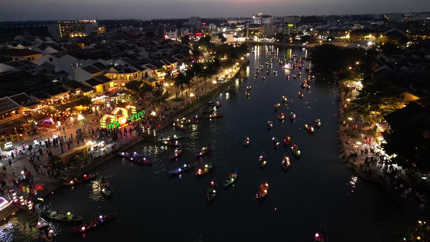 Aerial view of traditional lantern boats floating on the Hoai River at night with illuminated historic buildings in Hoi An, Da Nang, Vietnam.