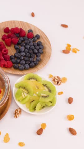 Hands place fresh kiwi slices into a bowl of cereal, berries, and nuts on white.