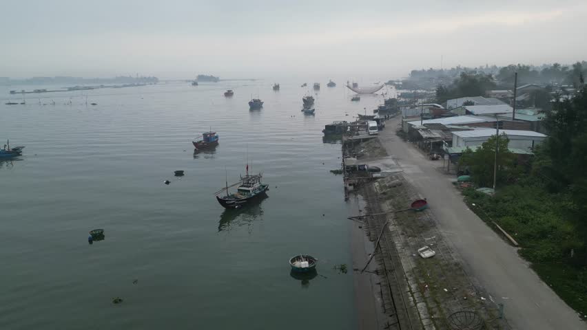 Aerial view of traditional fishing boats and round basket boats moored along the misty riverbank with local buildings and a coastal road in Hoi An, Da Nang, Vietnam.