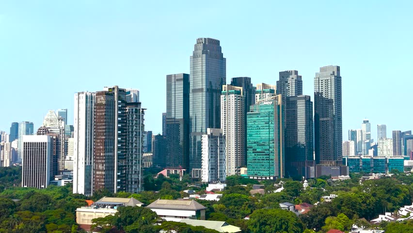 Aerial view of modern glass skyscrapers and high-rise buildings rising above lush green trees under a clear blue sky Jakarta, Jakarta, Indonesia.
