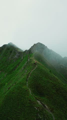 Aerial view of a narrow hiking trail winding along a steep green mountain ridge with mist rolling over the peaks and a hiker walking in Martigny, Valais, Switzerland.