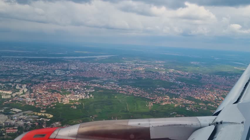 Aerial view of Belgrade, Serbia, from a passenger airplane on a cloudy day, featuring the winding Sava River and the vast urban sprawl of the Balkan capital beneath dramatic skies