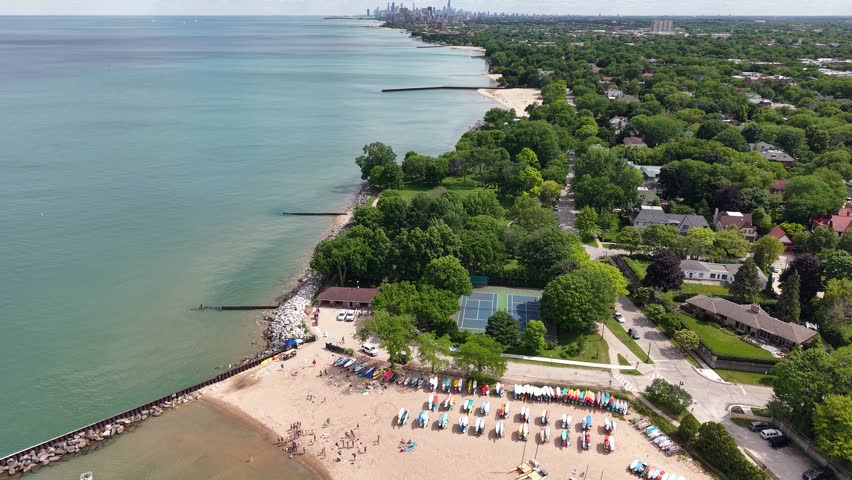 Aerial view of the Evanston, Illinois shoreline along Lake Michigan in summer, with the distant Chicago skyline on the horizon. Sailboats and paddleboards line the sandy beach beside turquoise water and lakefront neighborhoods. 4K UHD, 60fps, no audio.
