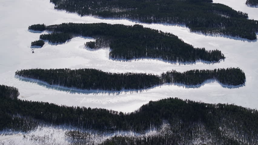 Aerial winter panorama of a frozen lake dotted with dark evergreen forest islands, forming winding archipelago shapes, icy channels across a vast boreal wilderness