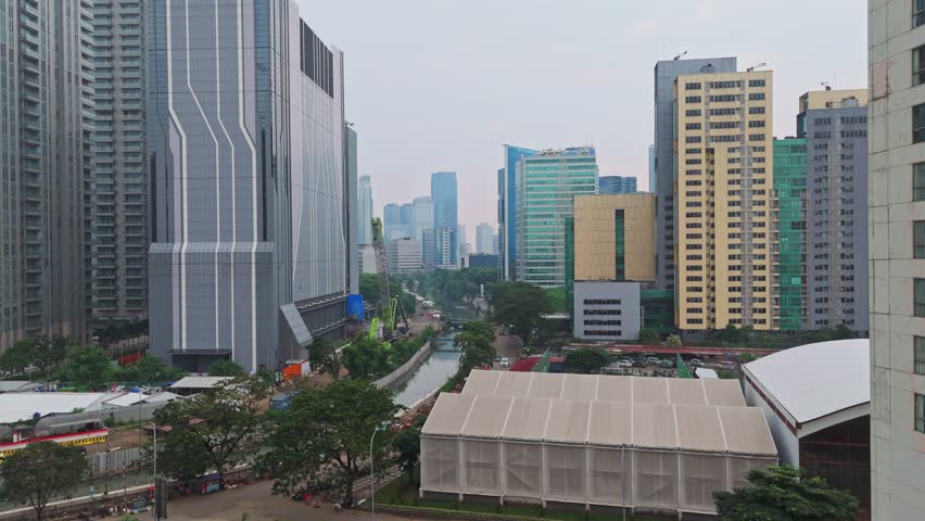 Drone aerial tilt-up shot of Jakarta cityscape with skyscrapers, river canal, and cloudy sky
