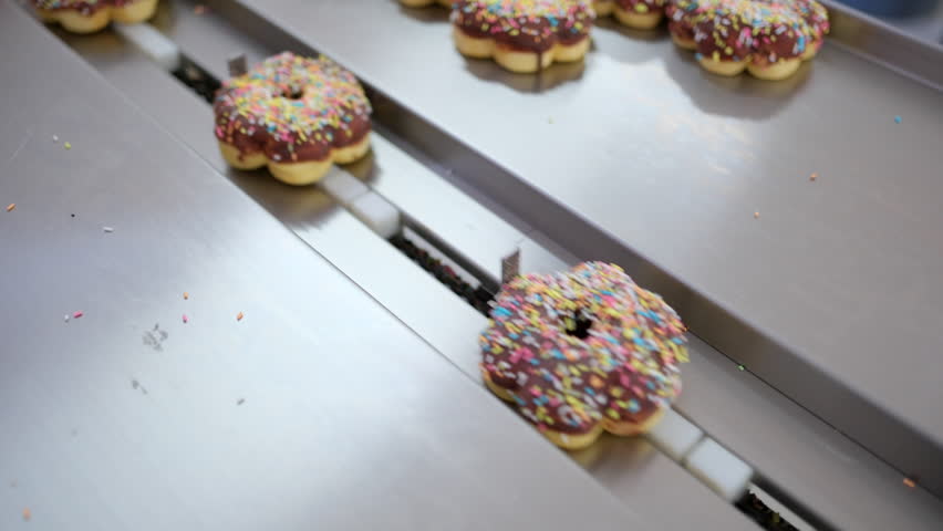 Freshly glazed donuts with colorful rainbow sprinkles travel along a stainless steel conveyor belt. Automated food production in a bakery