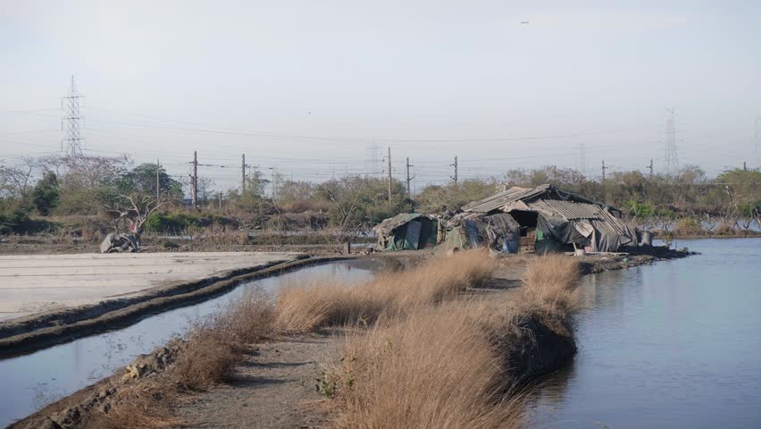 Daylight outdoors shot of a local or suburban train passing with salt lakes or pans filled with water and a makeshift hut in the foreground on a sunny windy day, Mumbai, India