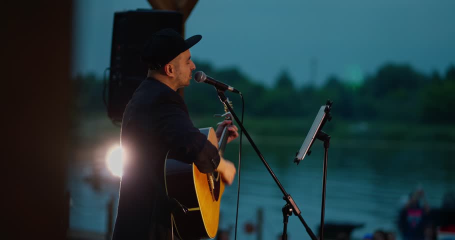 Male Acoustic Guitarist Singing on Riverside Wooden Stage at Dusk