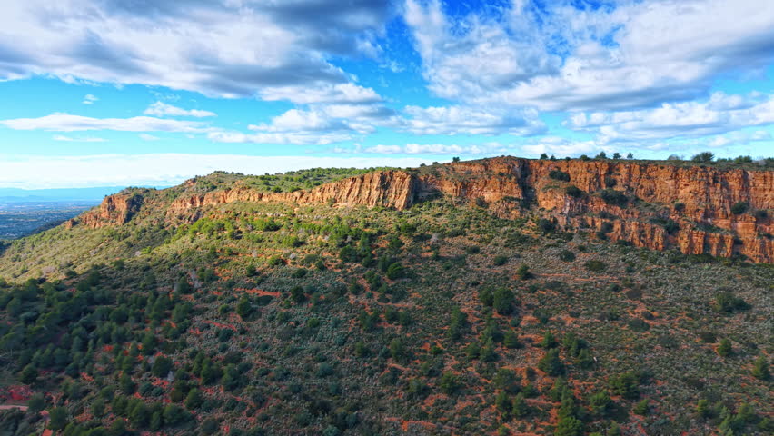 Bright day at rocky landscape in nature. Clouds float over a rocky hill filled with green plants and trees. The scene shows a vast area of land under a blue sky.