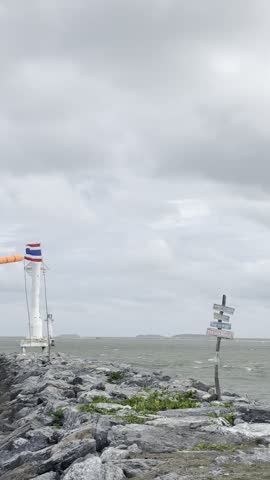 Small white lighthouse with Thai flag on a rocky breakwater under a dramatic stormy sky.