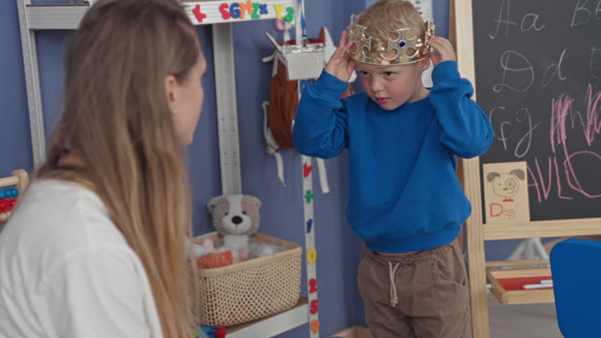 Woman teacher bending and guiding boy moving a blue chair by a chalkboard and height ruler inside a playroom building; curiosity.