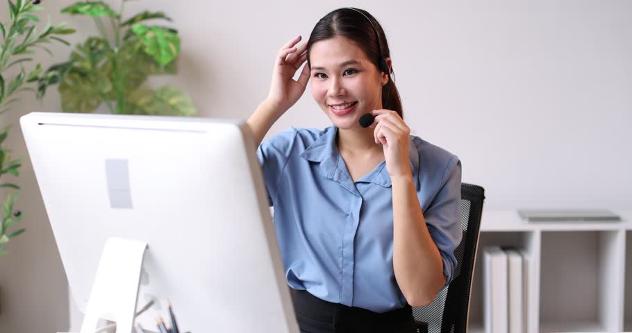 Friendly young asian woman working as a customer service representative, wearing a headset and smiling while providing support, assistance, and taking notes in a busy call center environment