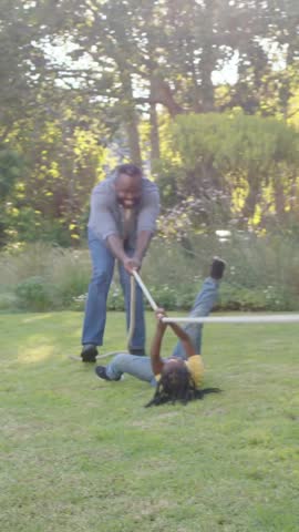 Vertical video: Pulling man in grey tugging rope, girl in yellow rising and pulling in yard playing. Family, garden, outdoor, kids, playful, laughter, vertical video